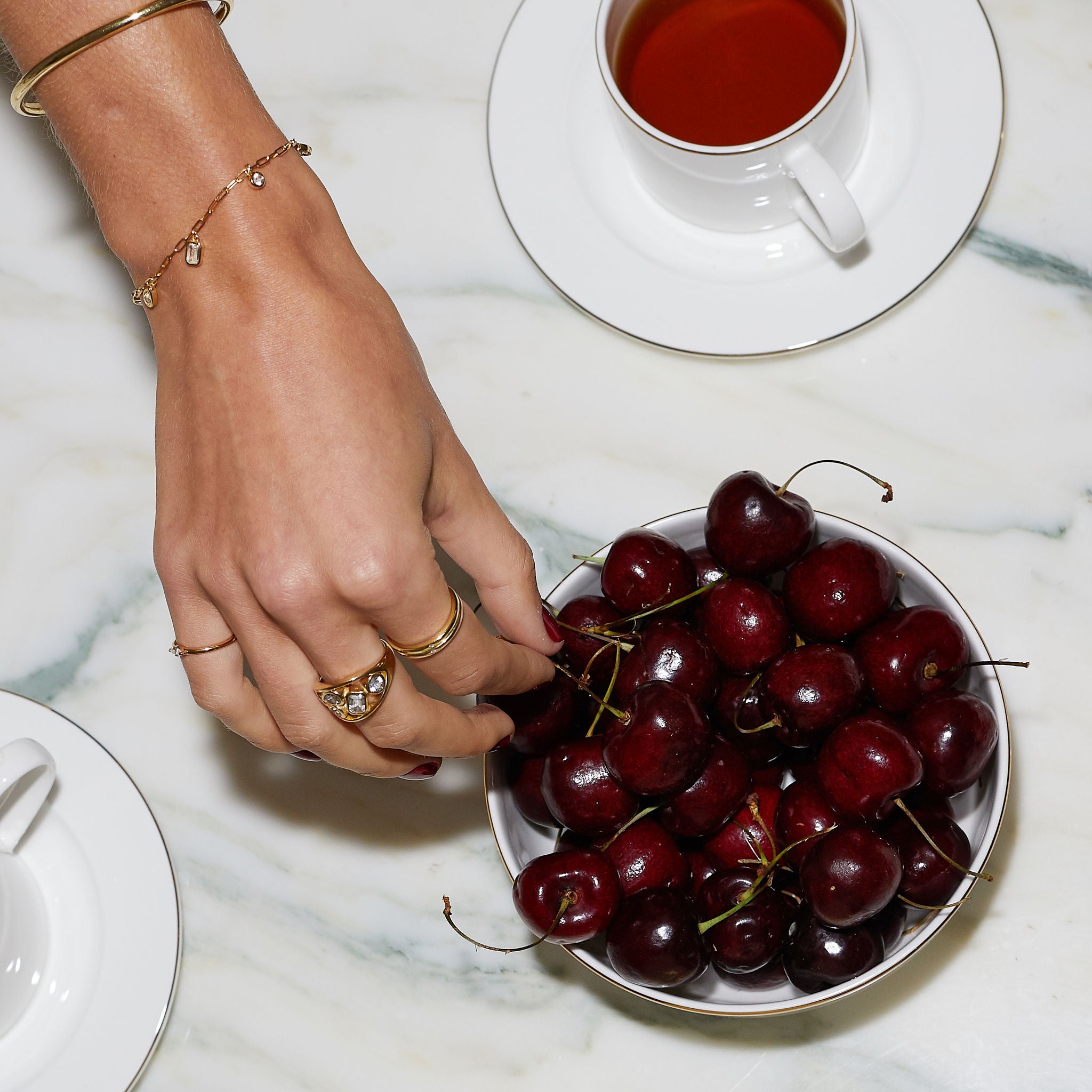 Person picking cherries from a bowl on a marble surface with a cup of tea in the background.