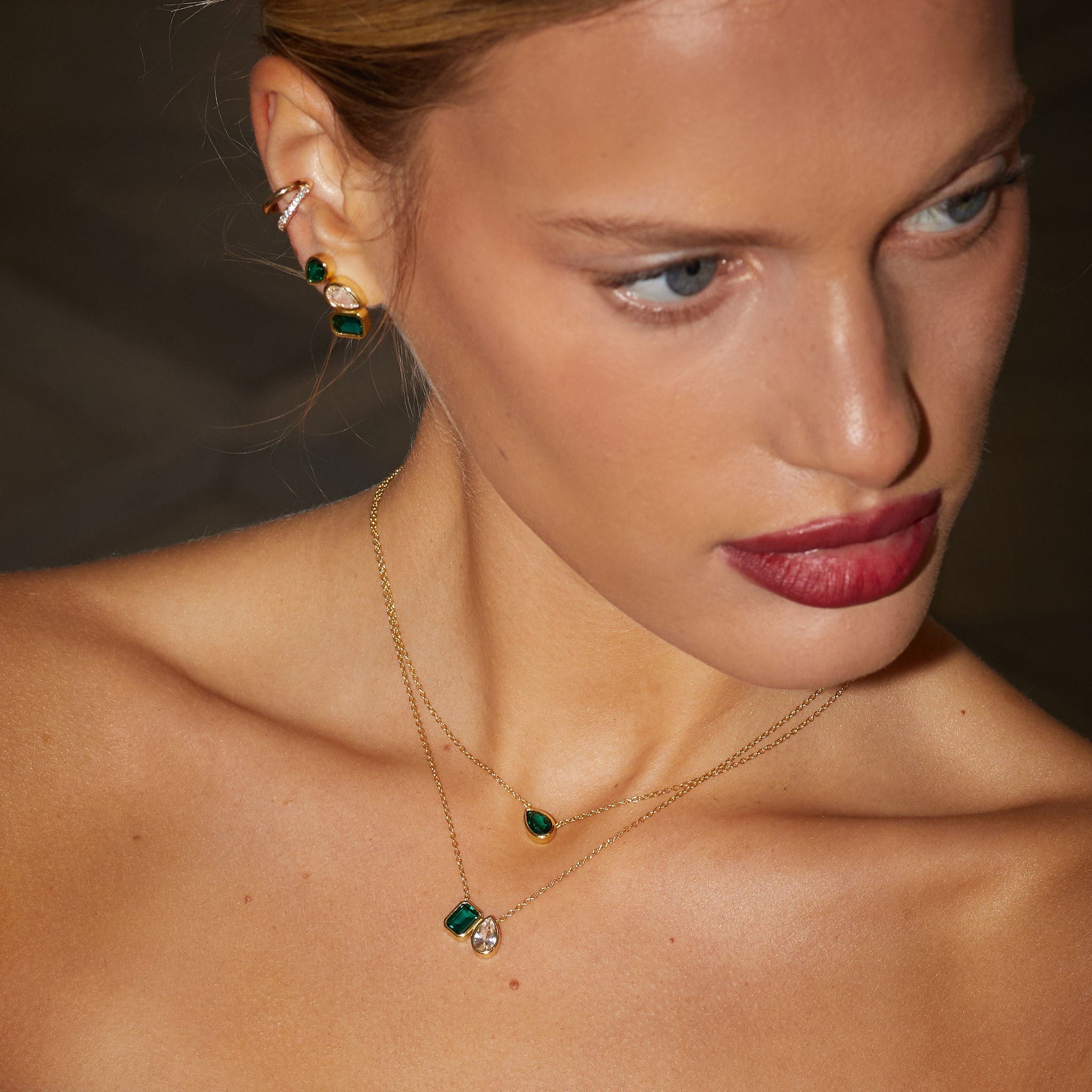 Woman wearing gold earrings and necklace with green gemstones against a dark background
