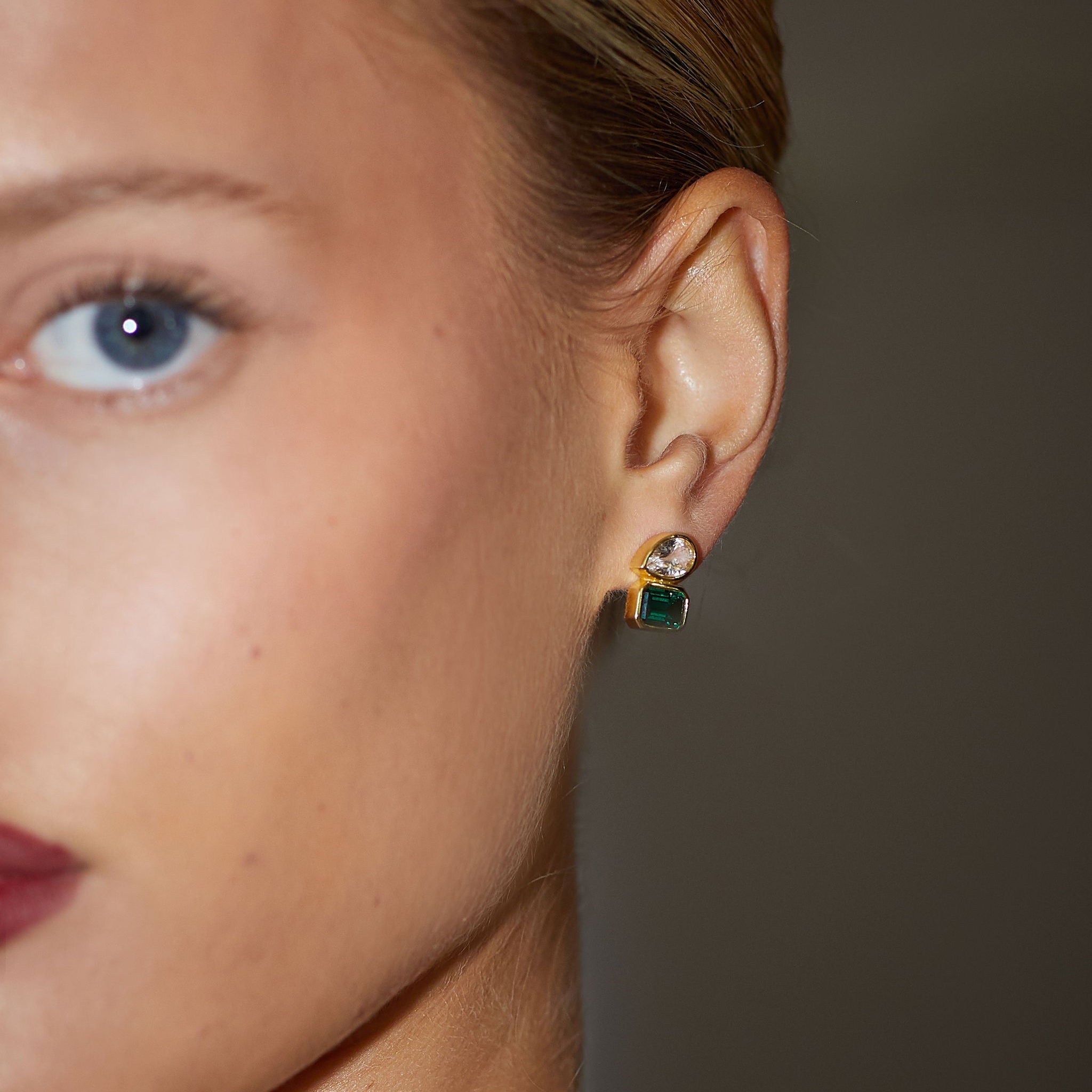 Close-up of a woman's ear wearing a diamond earring with a green gemstone against a neutral background