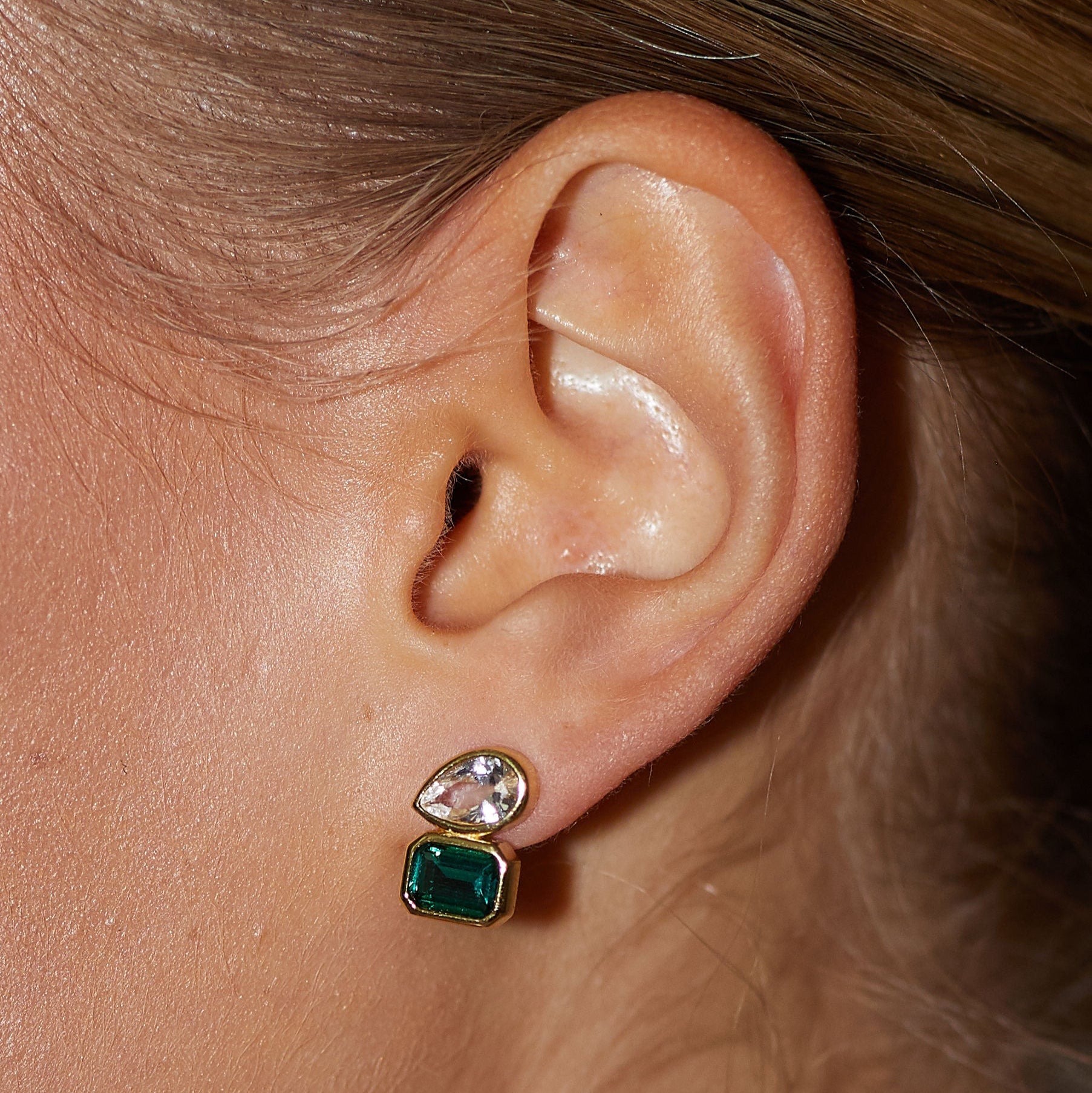 Close-up of an ear wearing a gold earring with green and clear gemstones.