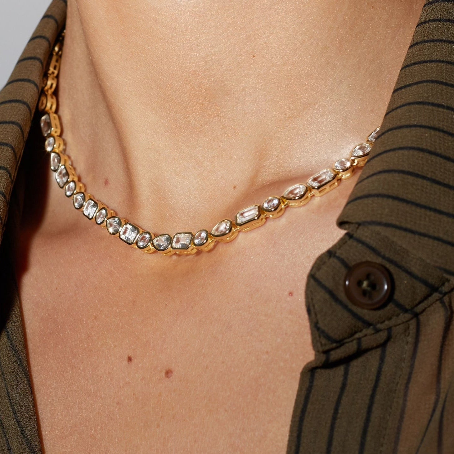 Gold necklace with clear stones worn by a person in a brown striped shirt.
